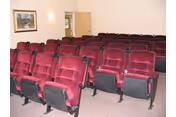 A small theater or presentation room with multiple rows of red cushioned seats arranged in front of a plain white wall. There is a framed picture on the left wall and an open door at the back of the room.