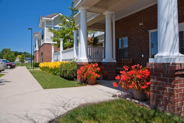 Exterior view of The Cortland Howell showing a brick building with white columns supporting a covered porch. There are vibrant red and yellow flowers in pots and flower beds along the walkway, with a clear blue sky in the background.