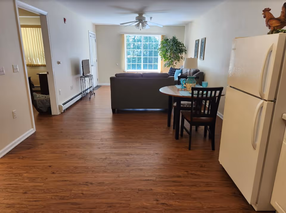 Interior view of a living space in an assisted living facility showing a wooden floor, a refrigerator on the right, a small round dining table with two chairs, a gray couch facing a window with vertical blinds, a TV on a stand, and a large potted plant near the window.