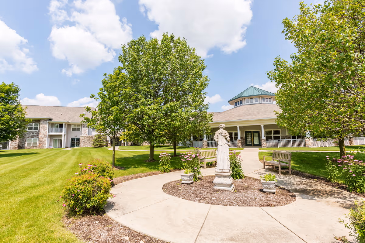 Outdoor garden area at Independence Village of Oxford (Waterstone) featuring a circular concrete pathway with a statue in the center, surrounded by green trees, bushes with pink flowers, wooden benches, and a building with a green roof and stone accents in the background under a partly cloudy blue sky.