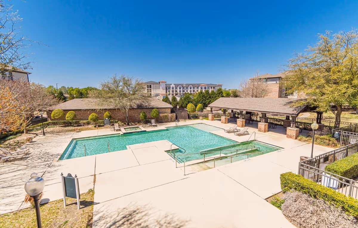 Outdoor swimming pool area with clear blue water surrounded by concrete decking. There are lounge chairs around the pool and a covered pavilion with brick pillars to the right. Trees and shrubs surround the pool area, and a multi-story building is visible in the background under a clear blue sky.