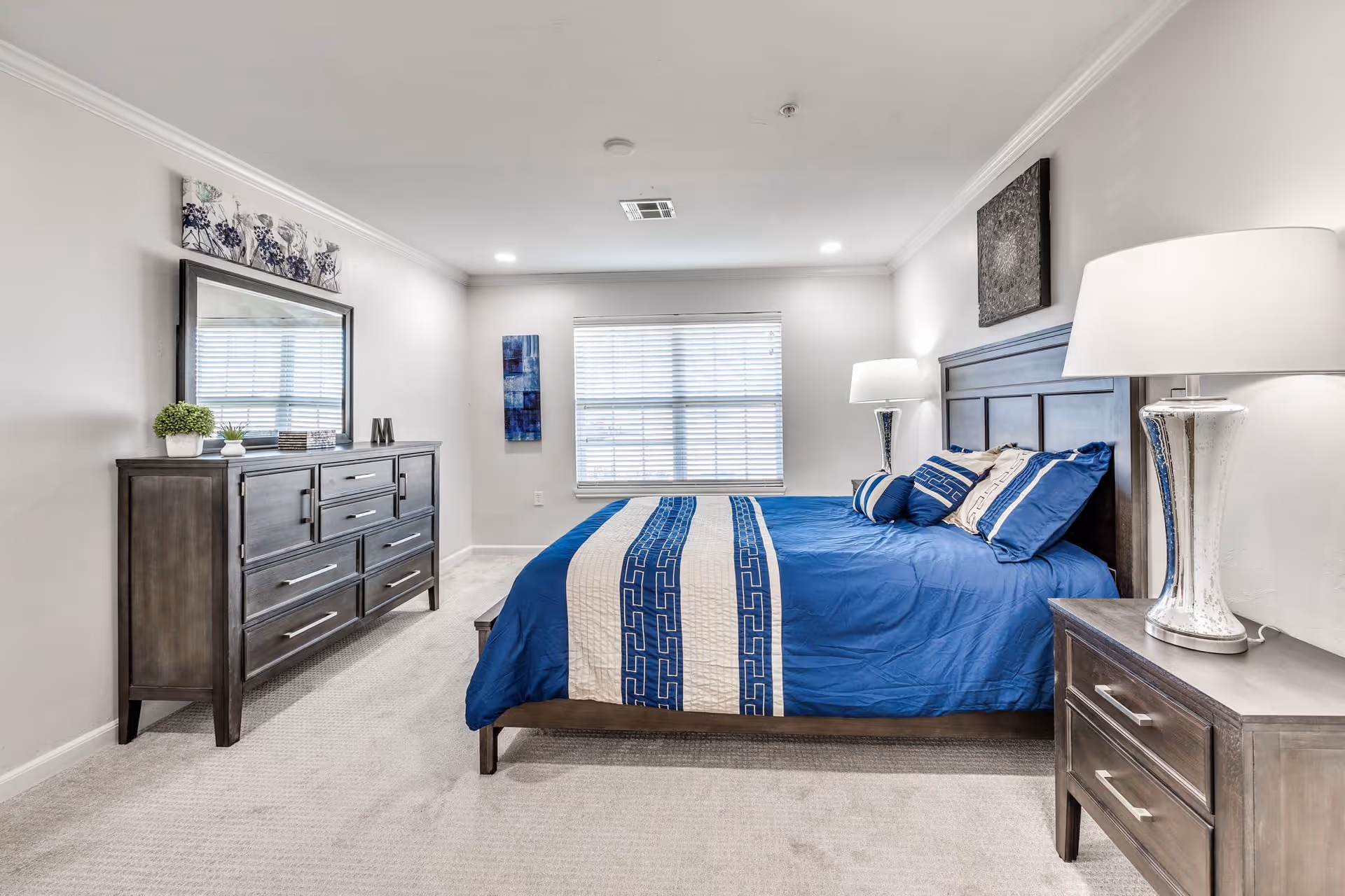 A bright and clean bedroom with a large bed featuring blue and white bedding. The room has a wooden headboard, matching wooden nightstands with lamps on each side, and a wooden dresser with a large mirror above it. There is a window with blinds letting in natural light, and the walls are decorated with modern artwork.