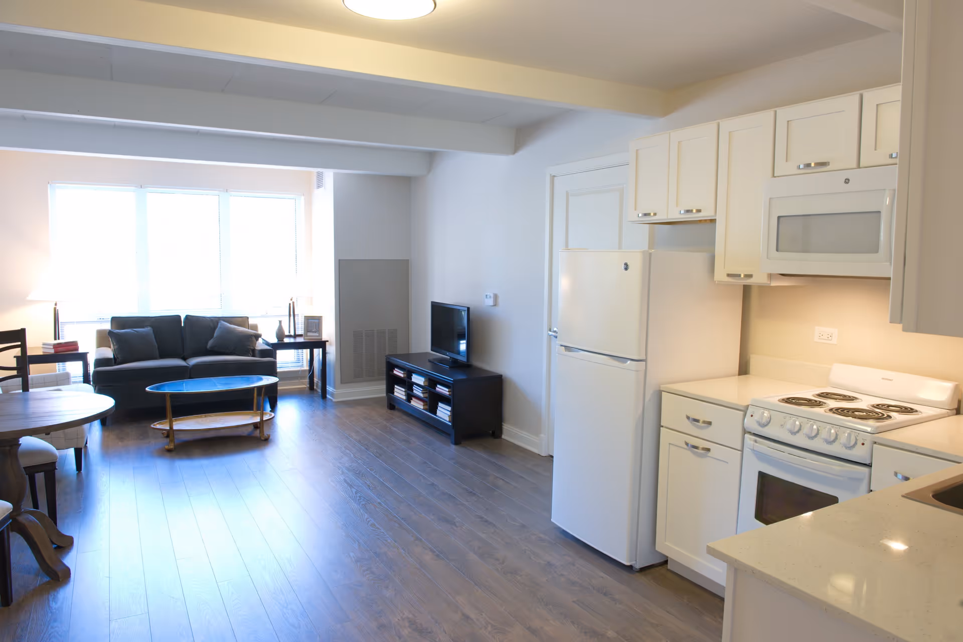 Bright open living space with a kitchen area on the right featuring white cabinets, a refrigerator, stove, and microwave. The living area on the left has a dark sofa with cushions, a coffee table, a TV on a stand, and a round dining table with chairs near a large window letting in natural light.