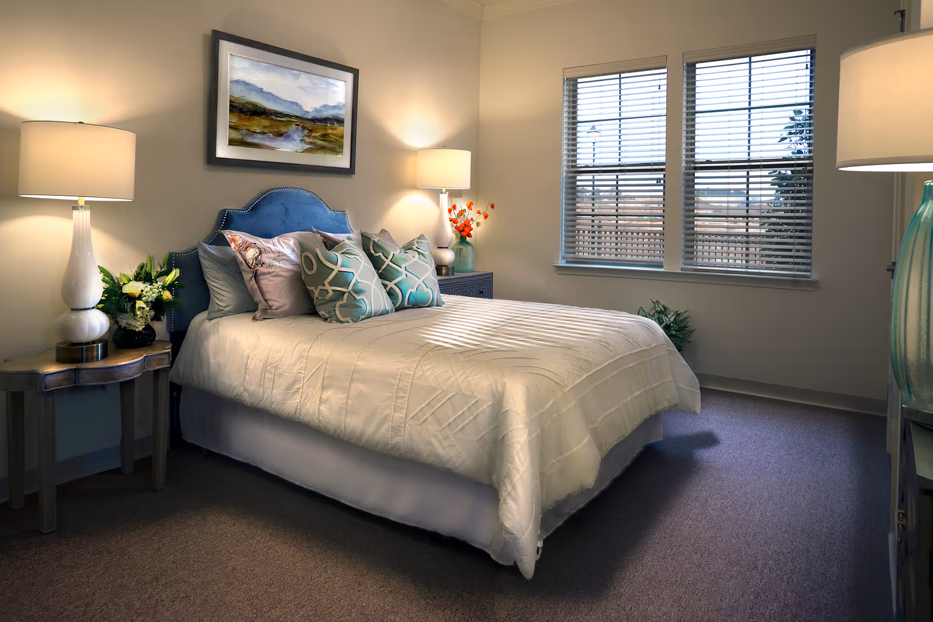 A cozy bedroom with a neatly made bed featuring a blue upholstered headboard and multiple decorative pillows. There are two bedside tables, each with a lamp and floral arrangements. A window with blinds allows natural light into the room, and a landscape painting hangs above the bed.