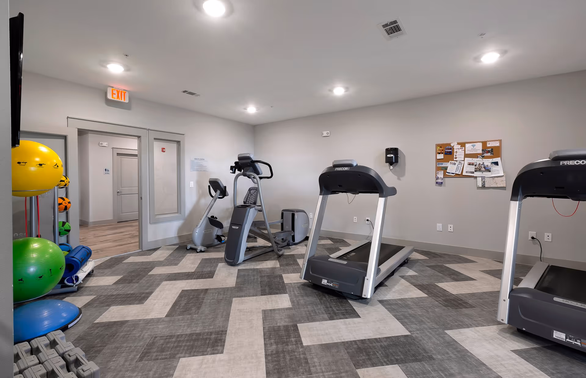 A fitness room with exercise equipment including two treadmills, an elliptical machine, and a stationary bike. There are colorful exercise balls and mats on a rack to the left. The room has a patterned floor, gray walls, ceiling lights, and a bulletin board with various notices on the right wall. An exit sign is visible above a door in the background.