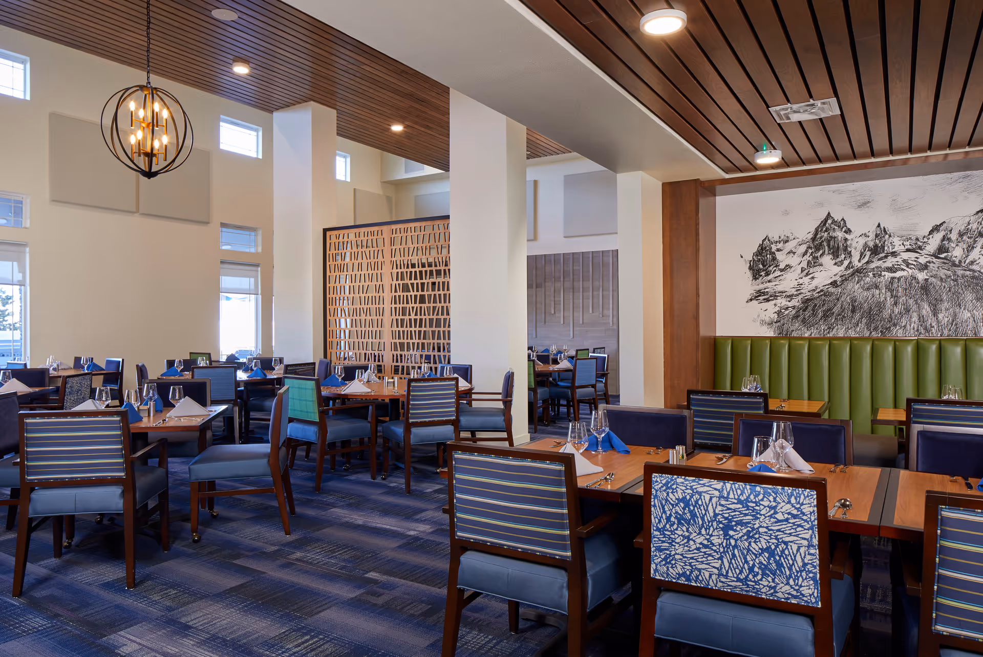 A spacious dining room with multiple wooden tables and chairs arranged neatly. The chairs have blue and patterned upholstery. Tables are set with wine glasses, napkins, and cutlery. The room features a blue carpet, white walls, a wooden slatted ceiling, and a decorative wooden partition. A large black and white mountain landscape artwork is displayed on one wall above a green cushioned bench.