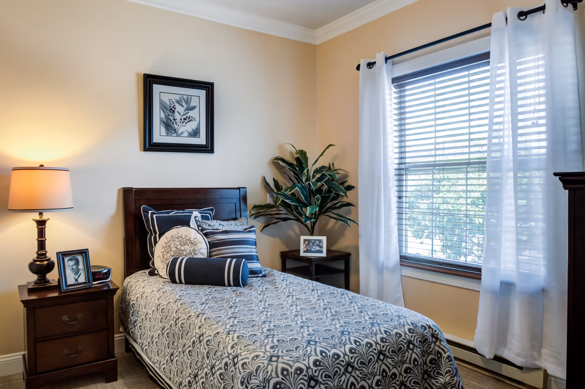 A cozy bedroom with a single bed featuring patterned bedding and multiple pillows. There is a wooden nightstand with a lamp and framed photo on the left side of the bed. A small table with a potted plant and another framed photo is positioned near a window with white curtains and blinds. A framed botanical print hangs on the beige wall above the bed.