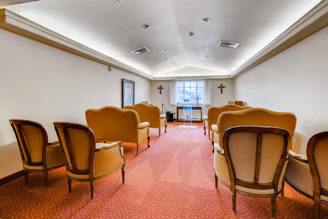 Interior view of a small chapel or meditation room with rows of vintage-style chairs and sofas arranged facing a podium in front of a window. The room has a red carpet with a dotted pattern, white walls, and two crosses mounted on either side of the window.