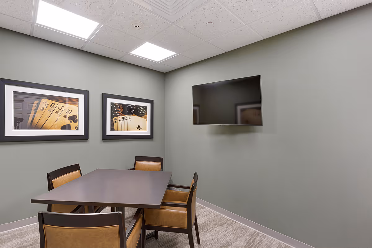 A small meeting or game room with a rectangular dark wood table surrounded by four brown leather chairs. The walls are painted light green and decorated with two framed pictures of playing cards. A flat-screen TV is mounted on the wall opposite the table. The room has a drop ceiling with fluorescent lighting.