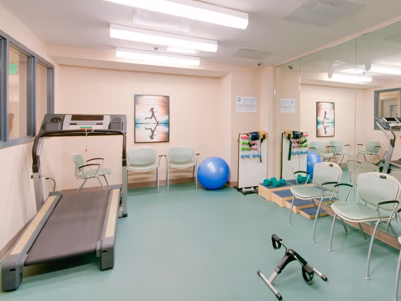 A small exercise room with a treadmill, several chairs, a large blue exercise ball, hand weights, resistance bands, and a pedal exerciser. The room has a large mirror on one wall and a framed picture of a person running on the opposite wall.