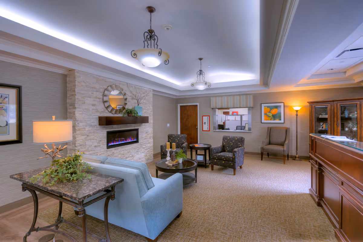 A cozy living room area in Woodhaven Retirement Community featuring a light blue sofa, two patterned armchairs, a stone fireplace with a modern electric fire, a round coffee table with candles and plants, a side table with a lamp and greenery, framed artwork on the walls, and warm lighting from ceiling fixtures and a floor lamp.