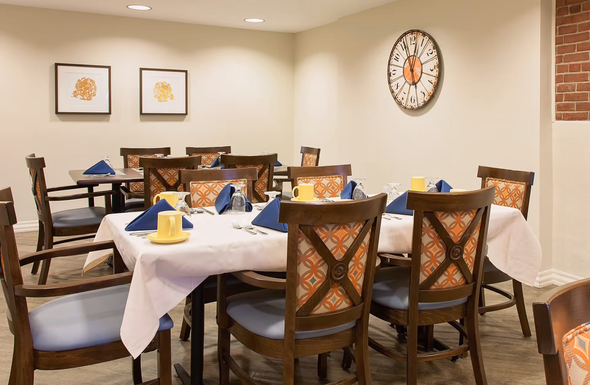 A dining room with several wooden tables and chairs. The chairs have patterned orange and beige cushions. Tables are set with white tablecloths, blue folded napkins, yellow mugs, upside-down glasses, and silverware. The walls are light-colored with two framed abstract artworks and a large round clock showing 11:55. The floor is wooden, and there is a partial view of a brick column on the right.