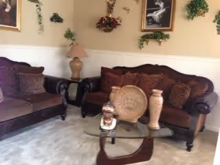 A cozy living room with two dark brown upholstered sofas adorned with multiple cushions. A glass coffee table with decorative vases and a small sculpture is placed in front of the sofas. The walls are decorated with framed pictures and green foliage accents, and a table lamp is positioned on a side table between the sofas.