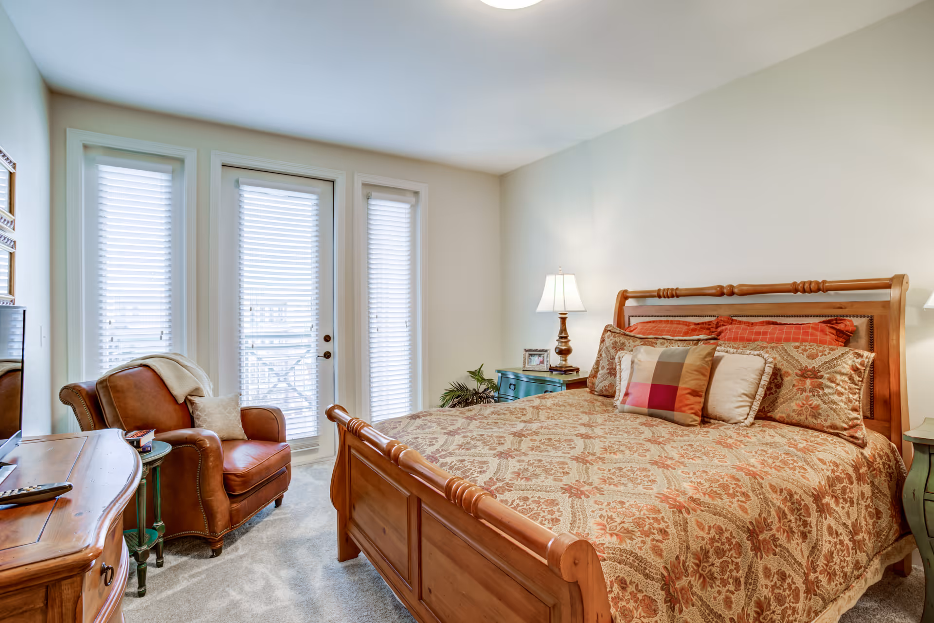 A cozy bedroom with a wooden bed frame and a patterned bedspread with multiple pillows. There is a brown leather armchair with a blanket and pillow next to a small round side table. The room has three tall windows with blinds and a door leading outside. Two bedside tables with lamps and a dresser with a TV are also visible.