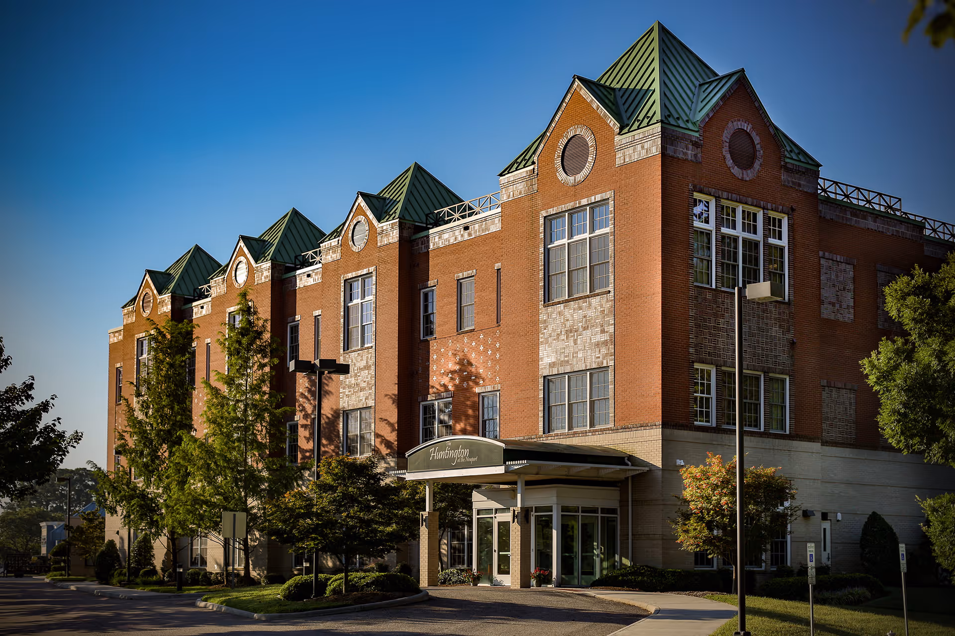 Exterior view of a multi-story brick building with green peaked roofs and large windows, surrounded by trees and landscaping under a clear blue sky. The entrance has a canopy with the sign 'Huntington Assisted Living'.