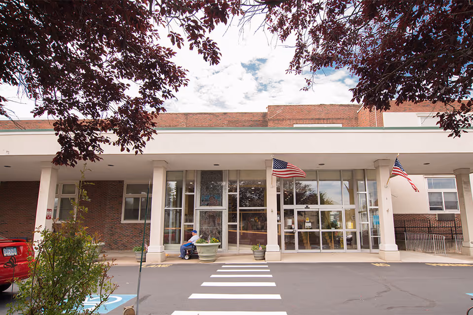 Front entrance of Saucon Valley Manor, a brick building with large glass doors and windows, two American flags mounted on the pillars, a person sitting on a bench near a large planter, and a crosswalk leading to the entrance.