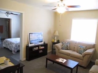 A cozy living room with a beige couch adorned with patterned pillows, a wooden coffee table with magazines, a small black TV stand holding a flat-screen TV and books, a side table with a lamp, and a ceiling fan with lights. A doorway leads to a bedroom with a bed covered in a floral bedspread.