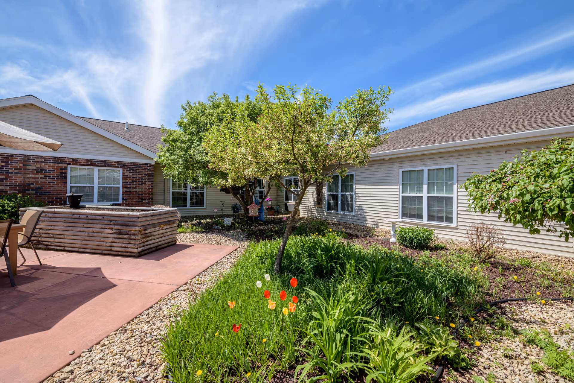 Outdoor garden area at Keystone Bluffs Assisted Living featuring a small tree, green plants, colorful flowers, and a patio with chairs and a wooden planter box under a bright blue sky with wispy clouds.