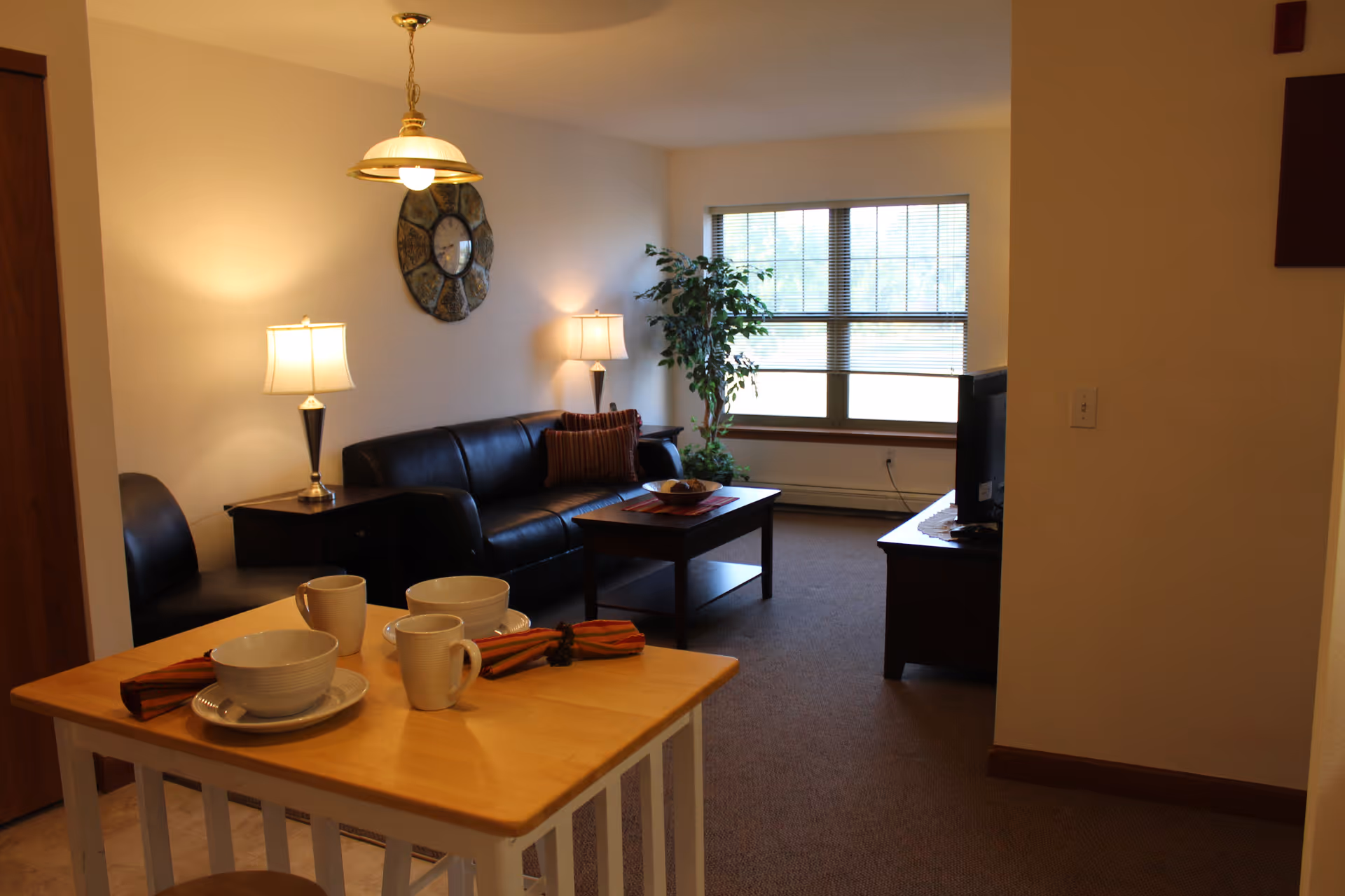 A cozy living room with a black leather sofa, coffee table, lamps, TV, and a small kitchen counter set with dishes.