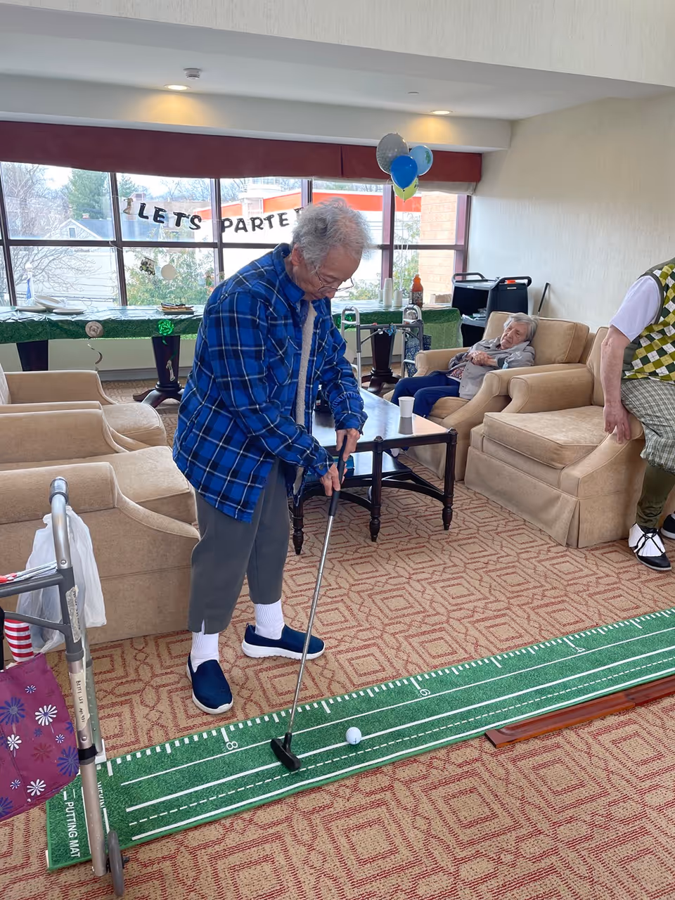 An elderly man in a blue plaid shirt playing indoor mini-golf on a putting mat in a living room area with beige armchairs and a coffee table. Another elderly person is seated on an armchair in the background, and a walker with a purple bag is visible on the left. There are balloons and a 'LET'S PARTY' banner on the window behind them.