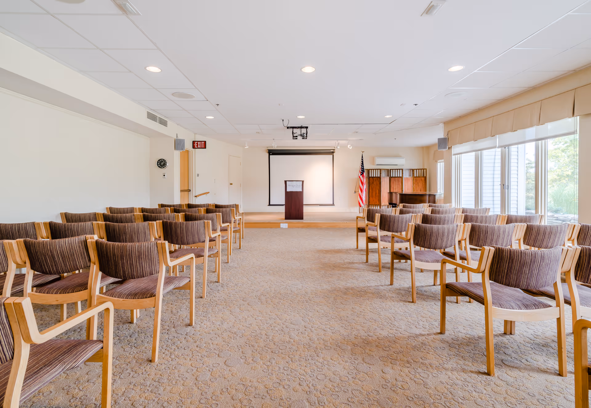 A conference or meeting room with rows of wooden chairs with cushioned seats and backs arranged facing a small stage with a podium and a projection screen. An American flag stands to the right of the stage, and large windows with blinds allow natural light into the room. The room has a beige carpet and white walls with ceiling lights.