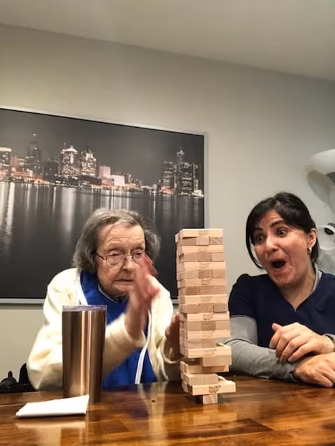 An elderly woman and a younger woman sitting at a wooden table playing a game of Jenga. The elderly woman is in the process of removing a block from the tower, while the younger woman watches with an excited and surprised expression. Behind them is a large framed photo of a city skyline at night.