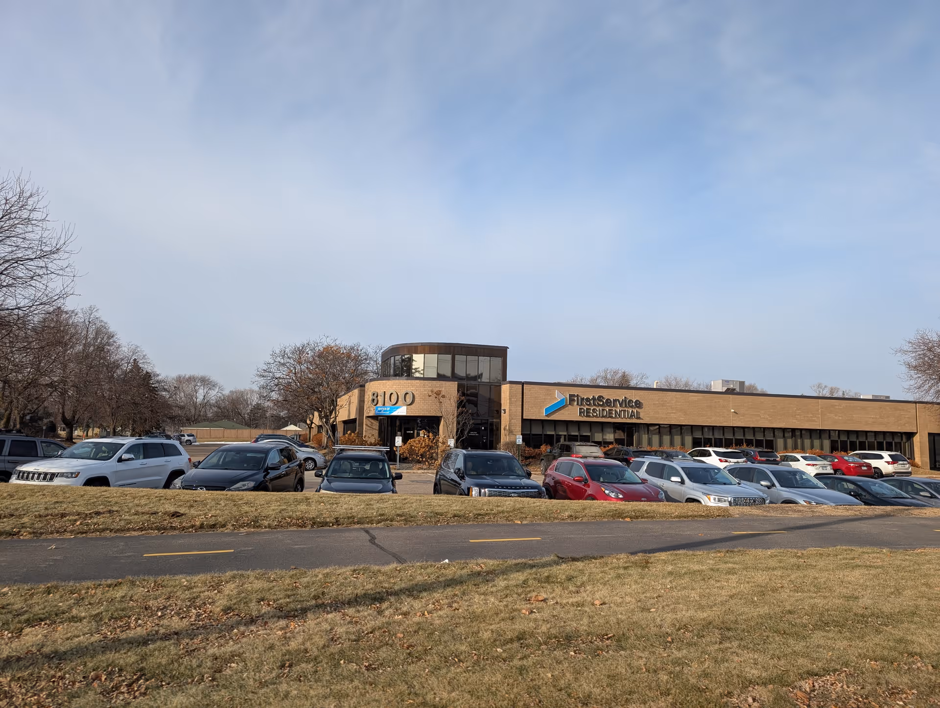 Exterior view of a single-story commercial building with a parking lot in front filled with cars. The building has large windows and a sign that reads 'FirstService Residential.' The address number 8100 is visible near the entrance. The sky is clear with some light clouds, and there is grass and a paved path in the foreground.