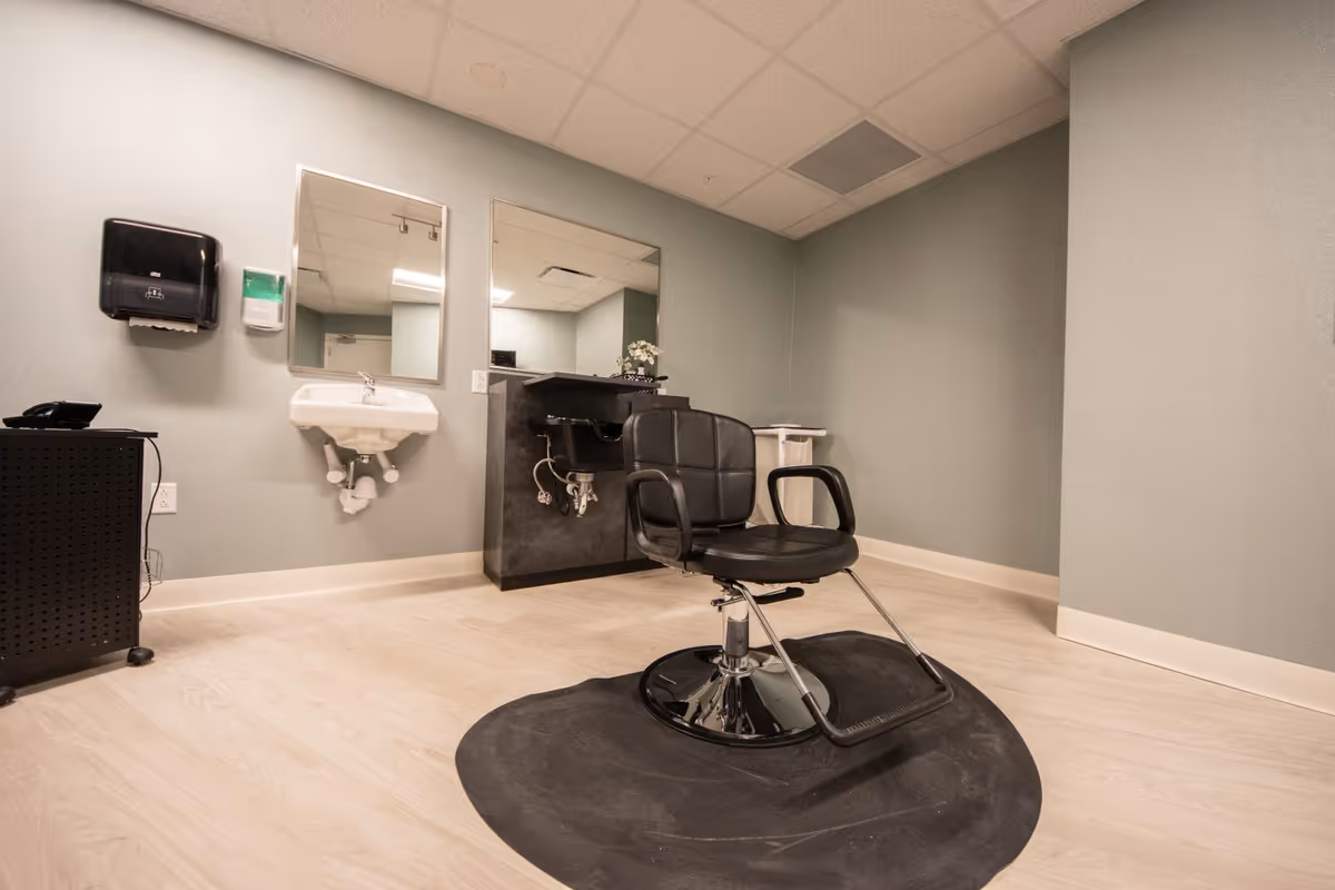 A small salon or barber room with a black salon chair on a black mat, a sink with a mirror above it, a black hair washing station with a mirror, a paper towel dispenser, and a soap dispenser mounted on the wall. The room has light wood flooring and light gray walls.