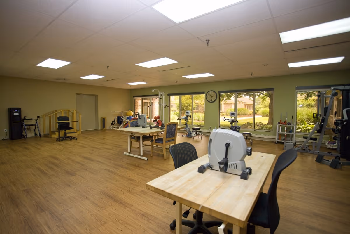A spacious rehabilitation or physical therapy room with wooden floors, several exercise machines, therapy tables, chairs, and large windows showing a garden outside. The room is well-lit with ceiling lights and has various equipment for physical therapy and exercise.