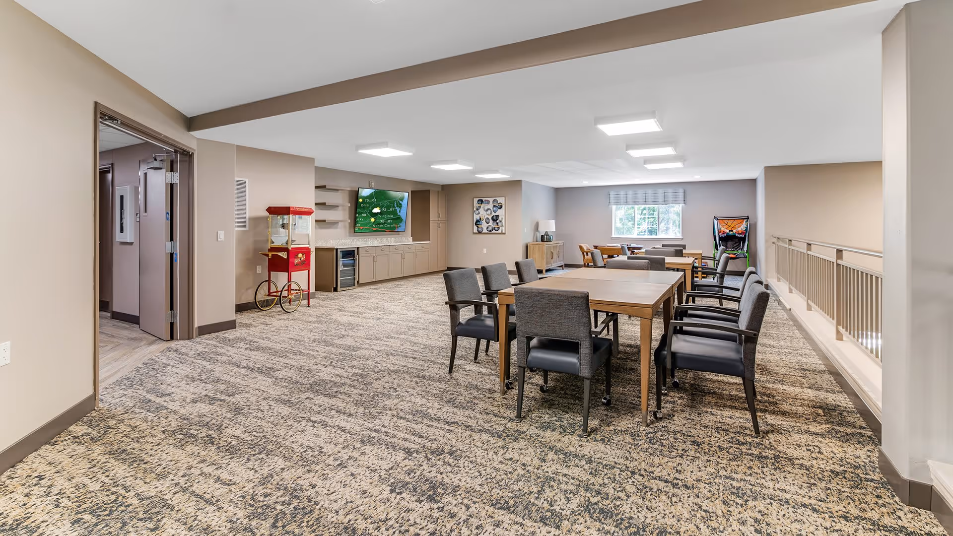 A spacious common room with carpeted floor, featuring several tables and chairs arranged for group seating. There is a popcorn machine near the wall, a large flat-screen TV mounted above a long cabinet, and a window letting in natural light. The room has neutral-colored walls and ceiling lights, with a railing on one side overlooking a lower level.