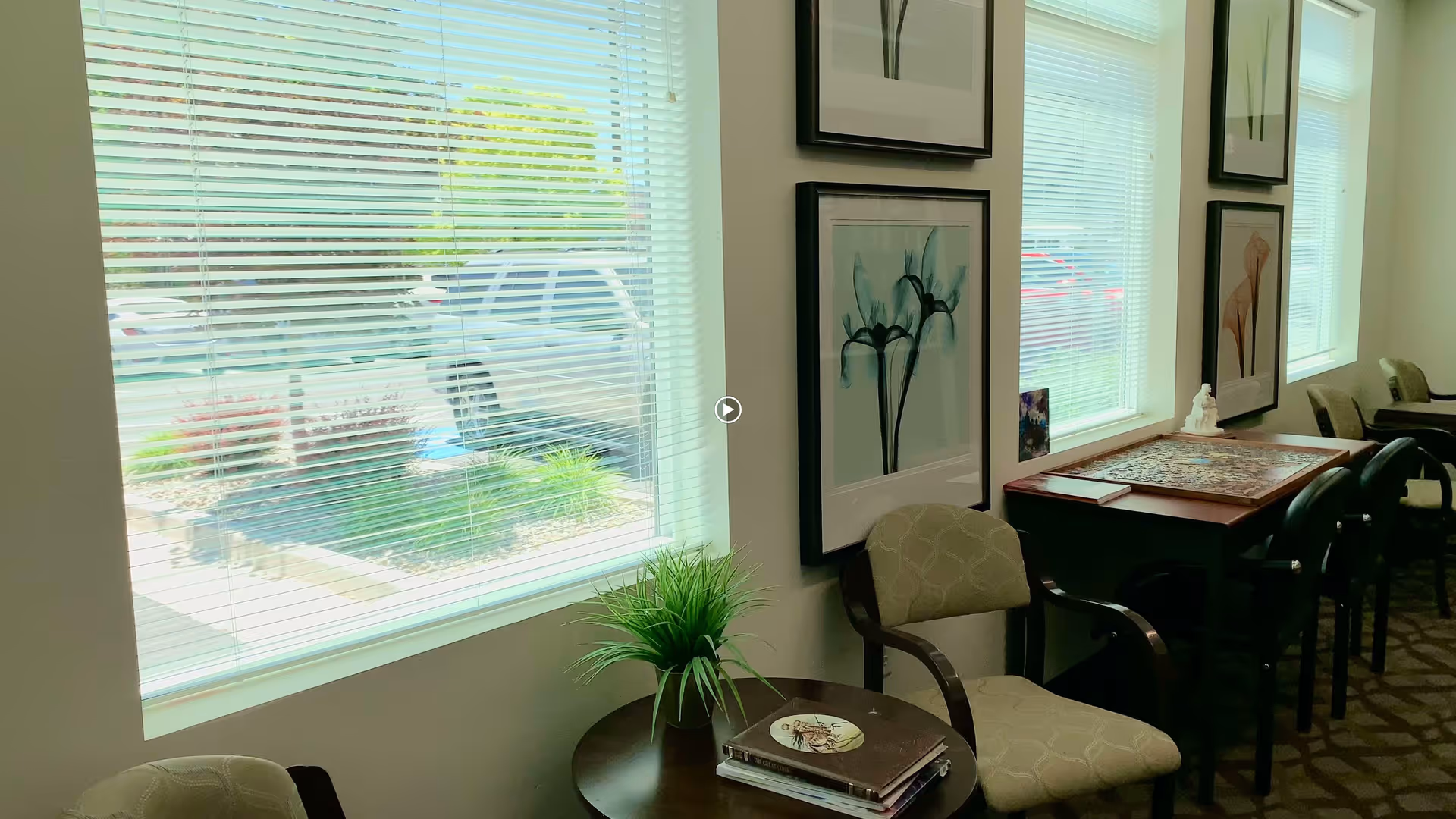 Interior view of a seating area with several chairs and tables next to large windows with blinds. The tables have decorative items including a plant and a puzzle. Framed floral artwork is hung on the wall above the chairs.