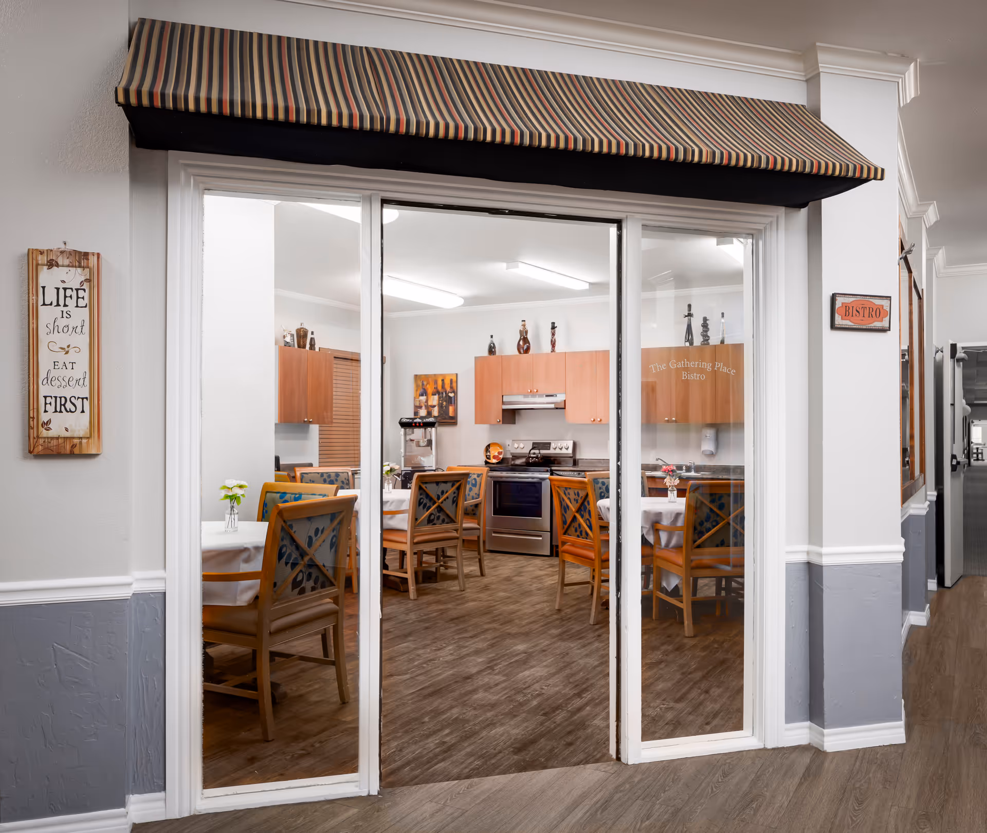View through glass sliding doors into a dining area with wooden chairs and tables covered with white tablecloths. The room has a kitchen area with wooden cabinets, a stove, and a coffee machine. A striped awning is above the doors, and signs on the walls read 'LIFE is short EAT dessert FIRST' and 'BISTRO'.