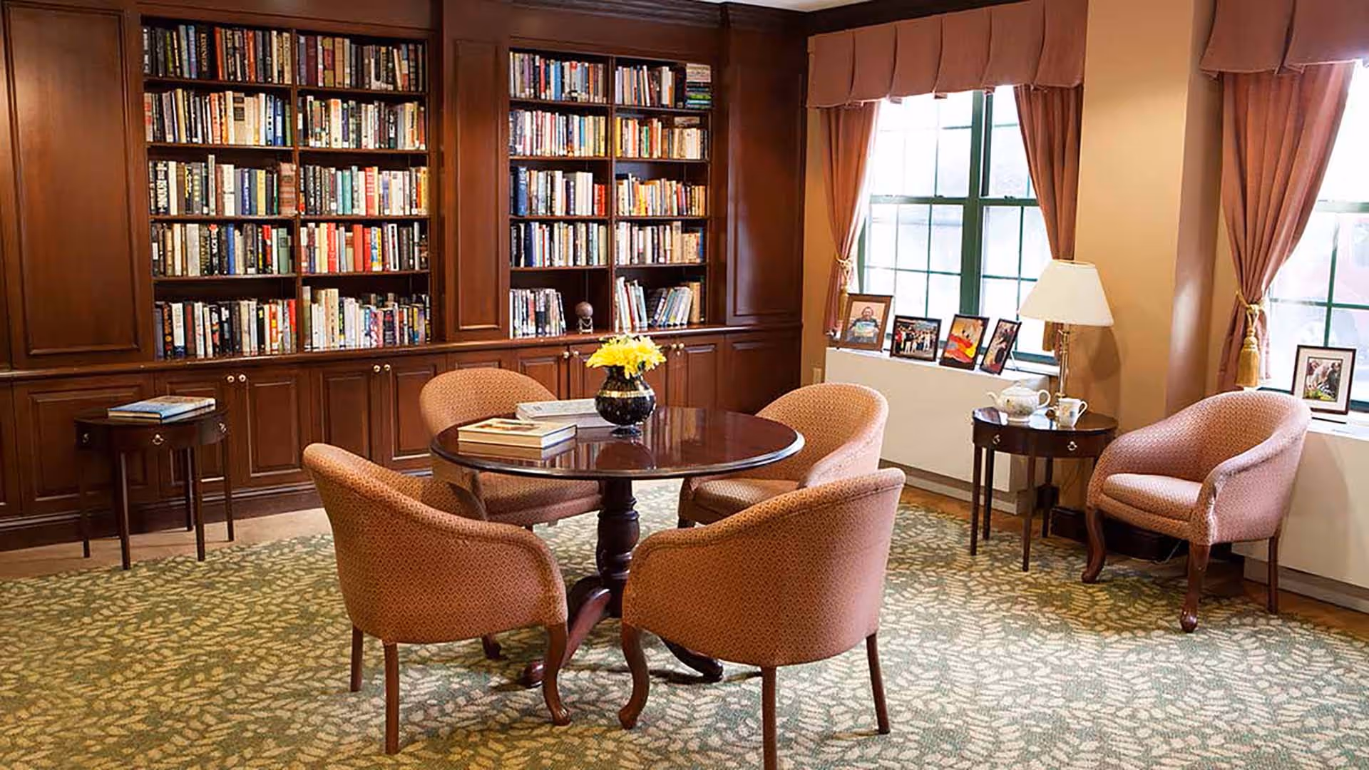 A cozy library room with wooden bookshelves filled with books, a round wooden table with a vase of yellow flowers and some books on it, surrounded by four upholstered chairs. There are two windows with brown curtains, a side table with a lamp and teapot, and framed photos on the windowsill.