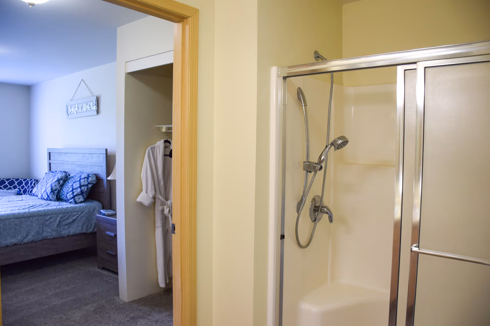 View of a bedroom with a bed, pillows, and a nightstand, seen through an open doorway from a bathroom area that features a shower with a sliding glass door and a handheld showerhead. A white bathrobe hangs in a small closet space between the bedroom and bathroom.