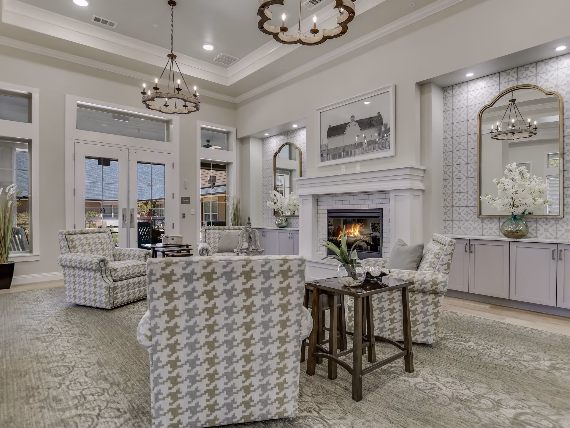 A cozy living room area in a senior living facility with four patterned armchairs arranged around a small wooden table. There is a lit fireplace with a white mantel, two large mirrors above built-in cabinets on either side, and a framed black and white picture above the fireplace. The room has large windows and glass doors letting in natural light, with two rustic chandeliers hanging from the ceiling.