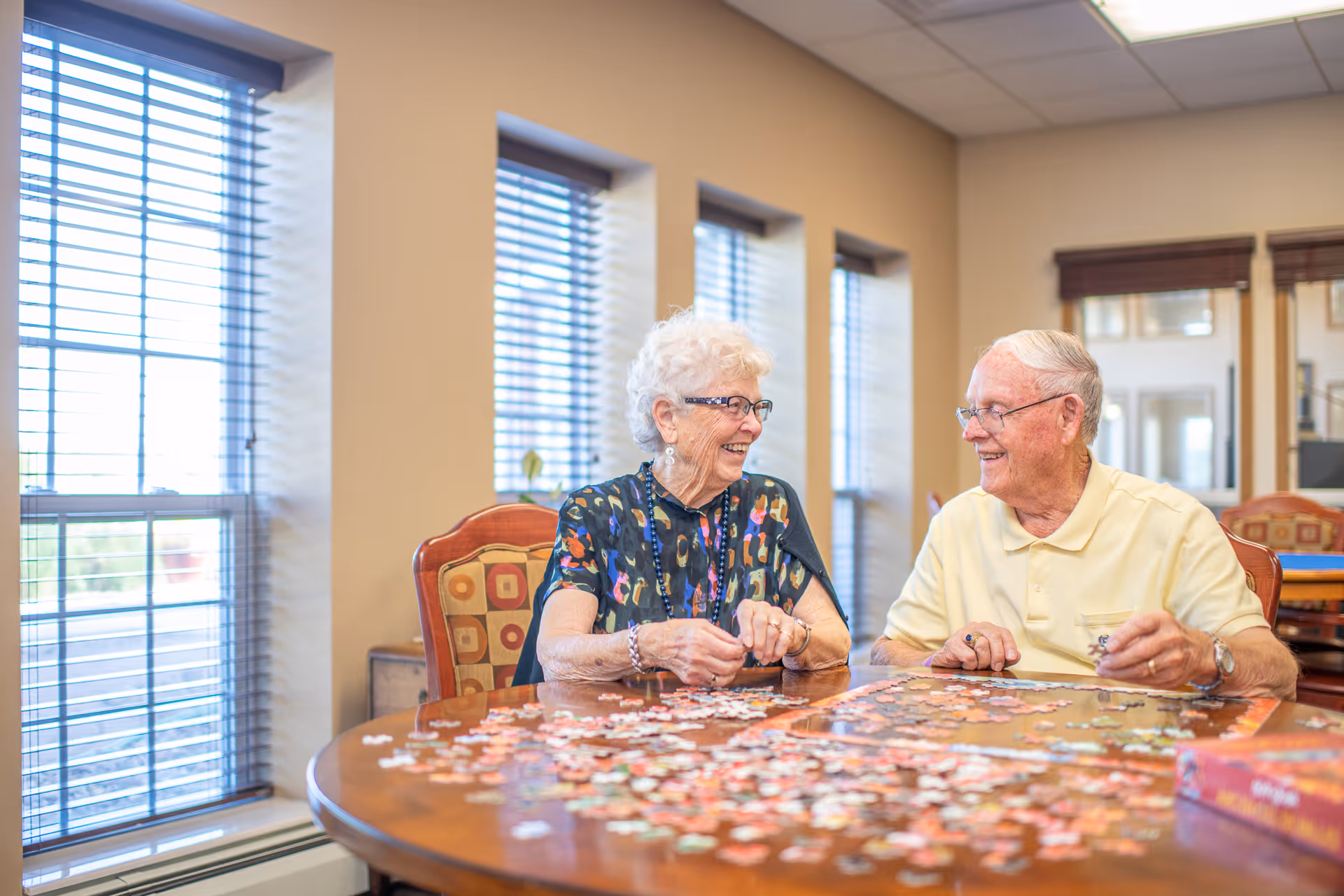 Two elderly individuals sitting at a round wooden table working on a jigsaw puzzle together in a well-lit room with large windows and beige walls.