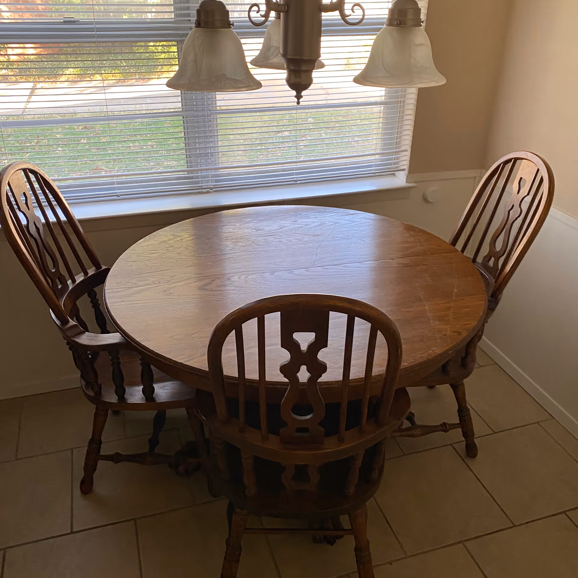 Round wooden dining table with three matching chairs under a hanging light by a window with blinds.