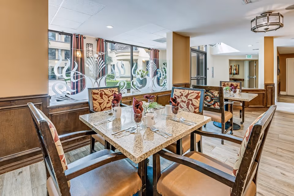 A dining area in a senior living facility with a square table set for four people. The table has granite top and is set with cups, silverware, and napkins folded in glasses. The chairs have floral patterned upholstery. Large windows with decorative frosted glass designs allow natural light to enter. The room has wood paneling and light-colored walls with a ceiling light fixture.