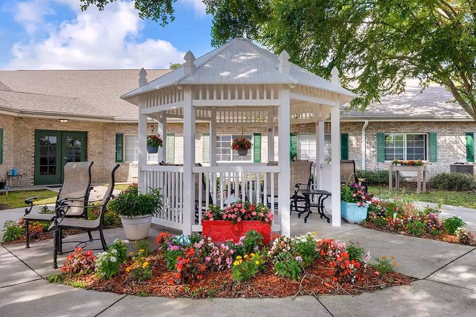 A white gazebo surrounded by colorful flower beds and potted plants in an outdoor garden area of a senior living facility. There are several chairs around the gazebo and a brick building with green shutters in the background under a partly cloudy sky.