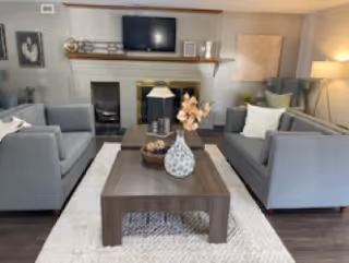 A cozy senior living room with two gray sofas facing each other across a wooden coffee table. The table holds decorative items including a vase with flowers. Behind the sofas is a white fireplace with a mounted flat-screen TV above it. The room is softly lit with a floor lamp and decorated with framed artwork and cushions.