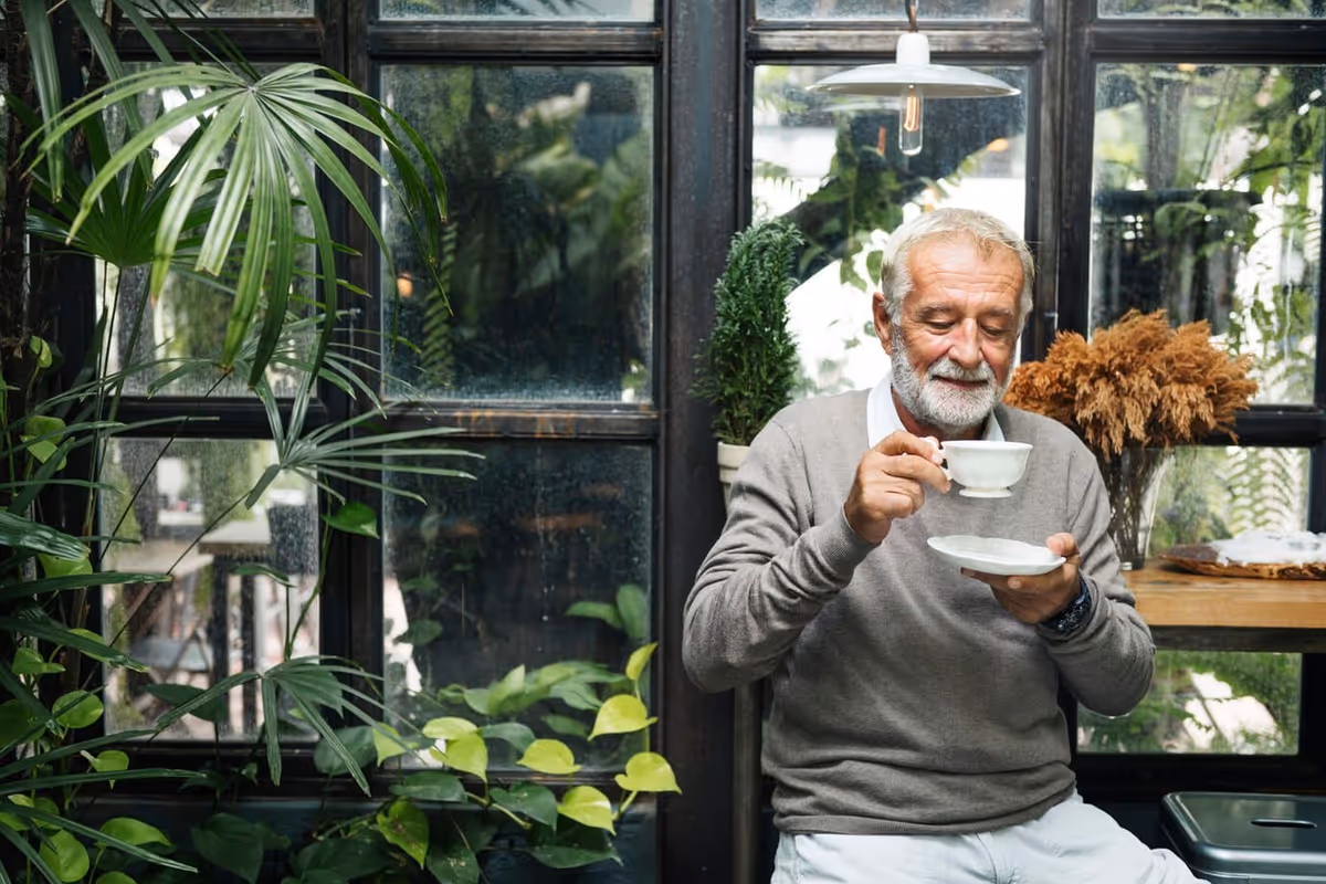 An elderly man with gray hair and a beard is sitting indoors near large windows surrounded by green plants. He is holding a white teacup and saucer, appearing to enjoy a moment of calm while drinking tea.