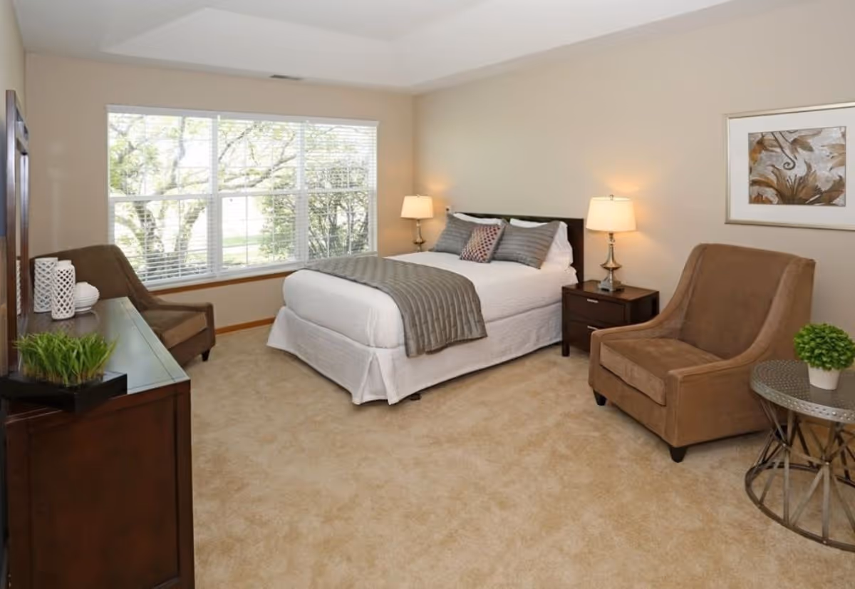A cozy bedroom with a large window letting in natural light. The room features a neatly made bed with white bedding and gray pillows, two bedside tables with lamps, a brown armchair next to a small round table with a potted plant, and a dresser with decorative items. The walls are painted beige and there is a framed artwork hanging on the wall.