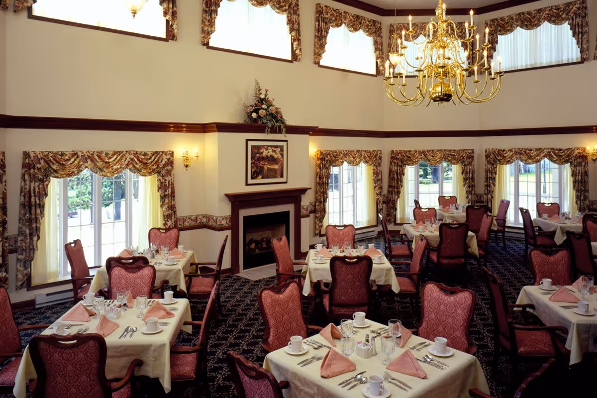 Elegant dining room with multiple tables set with white tablecloths, pink folded napkins, cups, glasses, and silverware. The room features large windows with floral curtains, a fireplace with a floral arrangement and painting above it, and a large gold chandelier hanging from the ceiling.