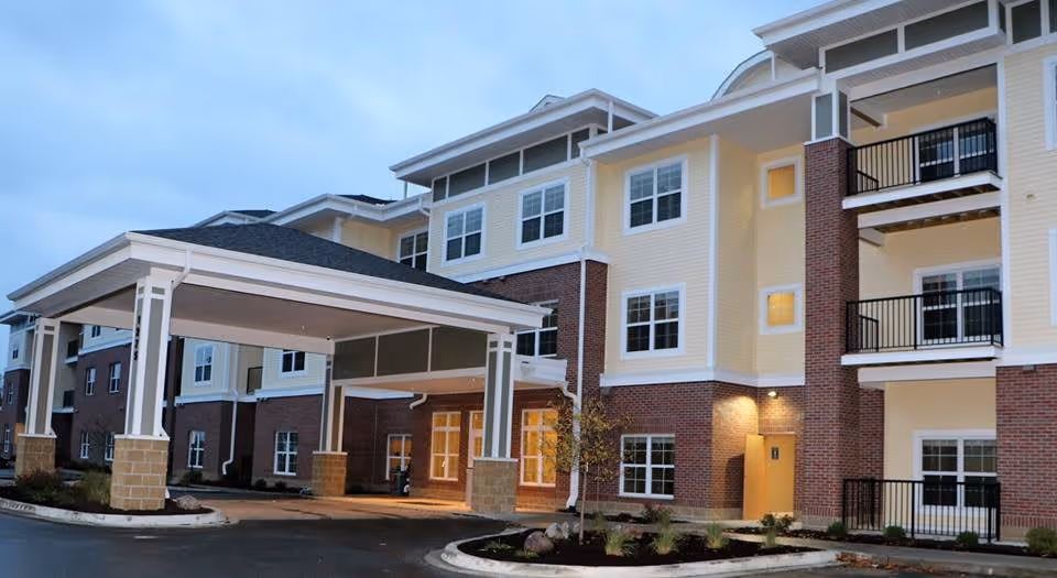Exterior view of a multi-story senior living facility building with a covered entrance, brick and light yellow siding, multiple windows, balconies, and landscaped surroundings during early evening.