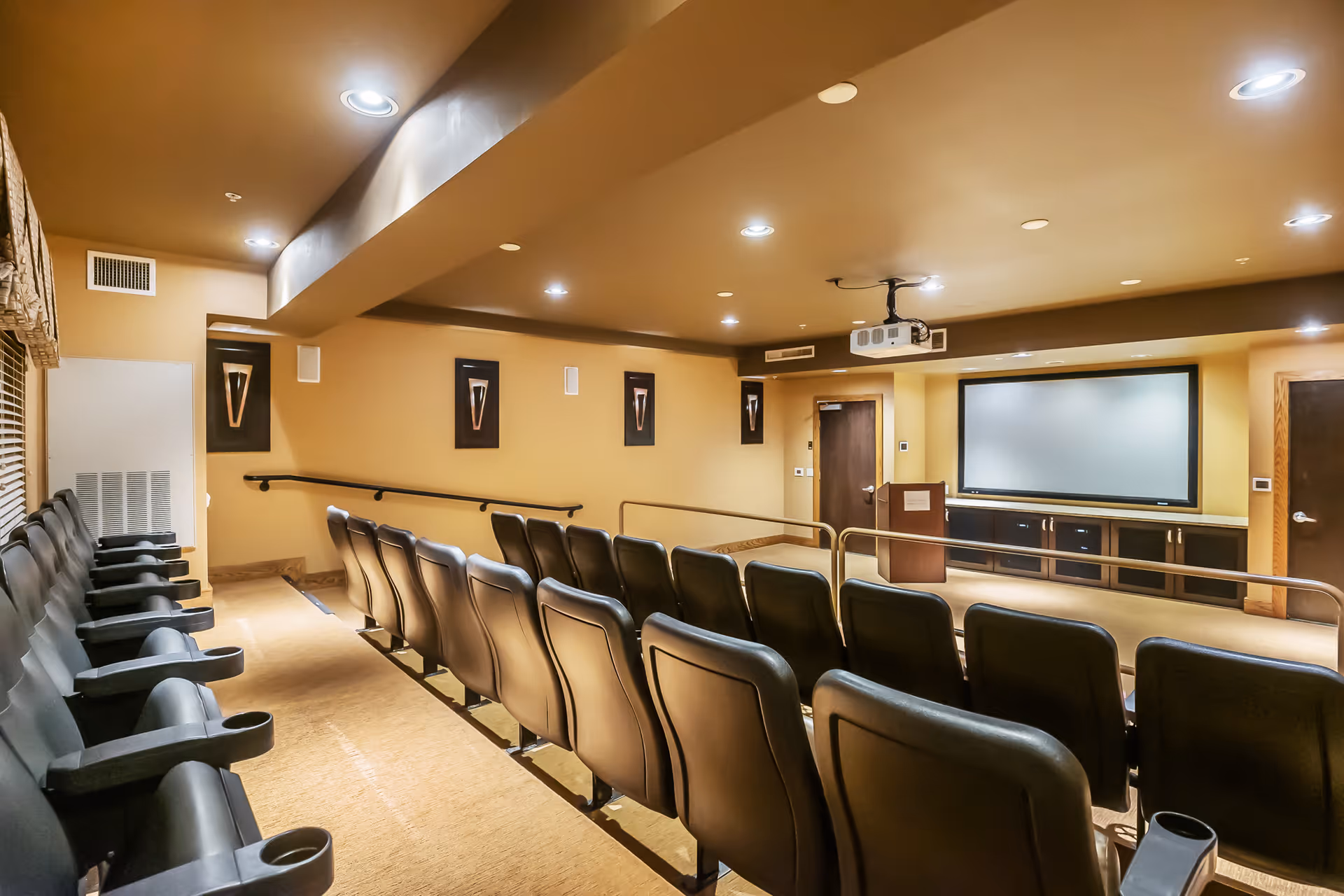 A small theater room with rows of black leather seats facing a large screen mounted on a beige wall. The room has a beige carpet and ceiling with recessed lighting, and there is a projector mounted on the ceiling. The walls are decorated with framed artwork, and there are handrails along the aisles.