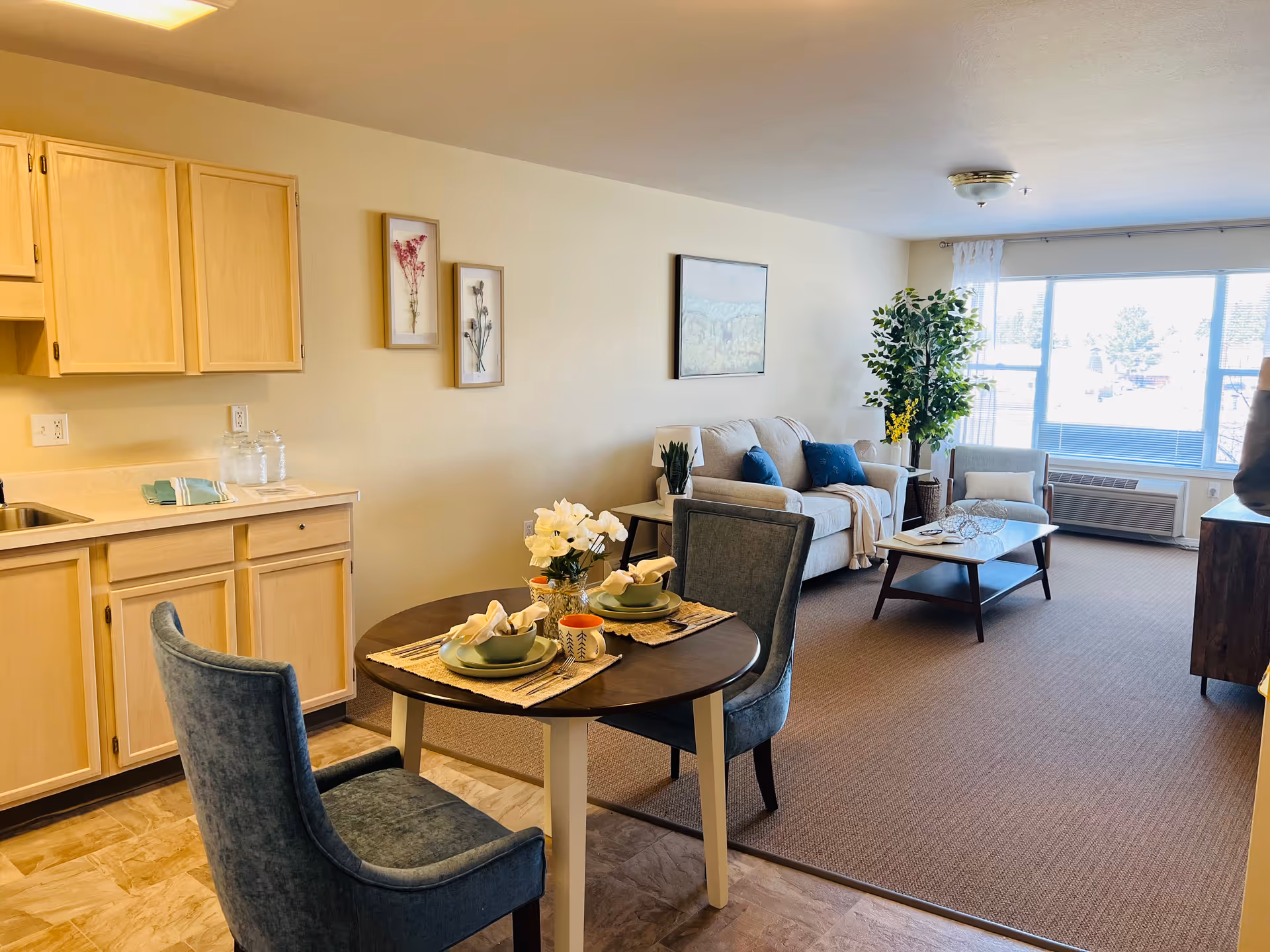 Interior view of a senior living facility apartment showing a small dining area with a round table set for two, a kitchenette with light wood cabinets, and a living room with a beige sofa, blue cushions, a coffee table, an armchair, a large window, and decorative plants and artwork on the walls.