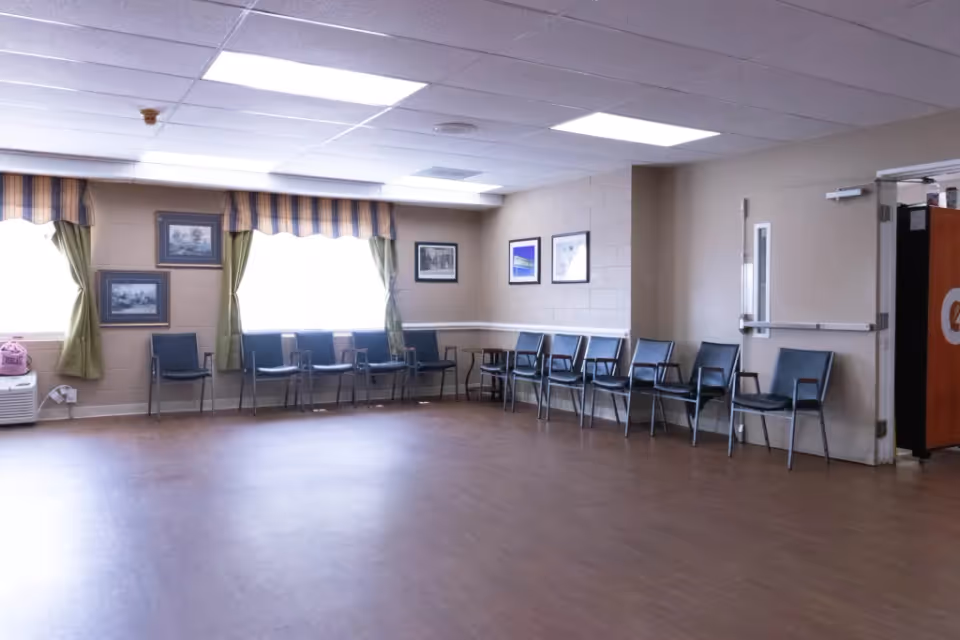 Empty common activity room with chairs lined along beige walls, windows with green curtains and framed artwork.