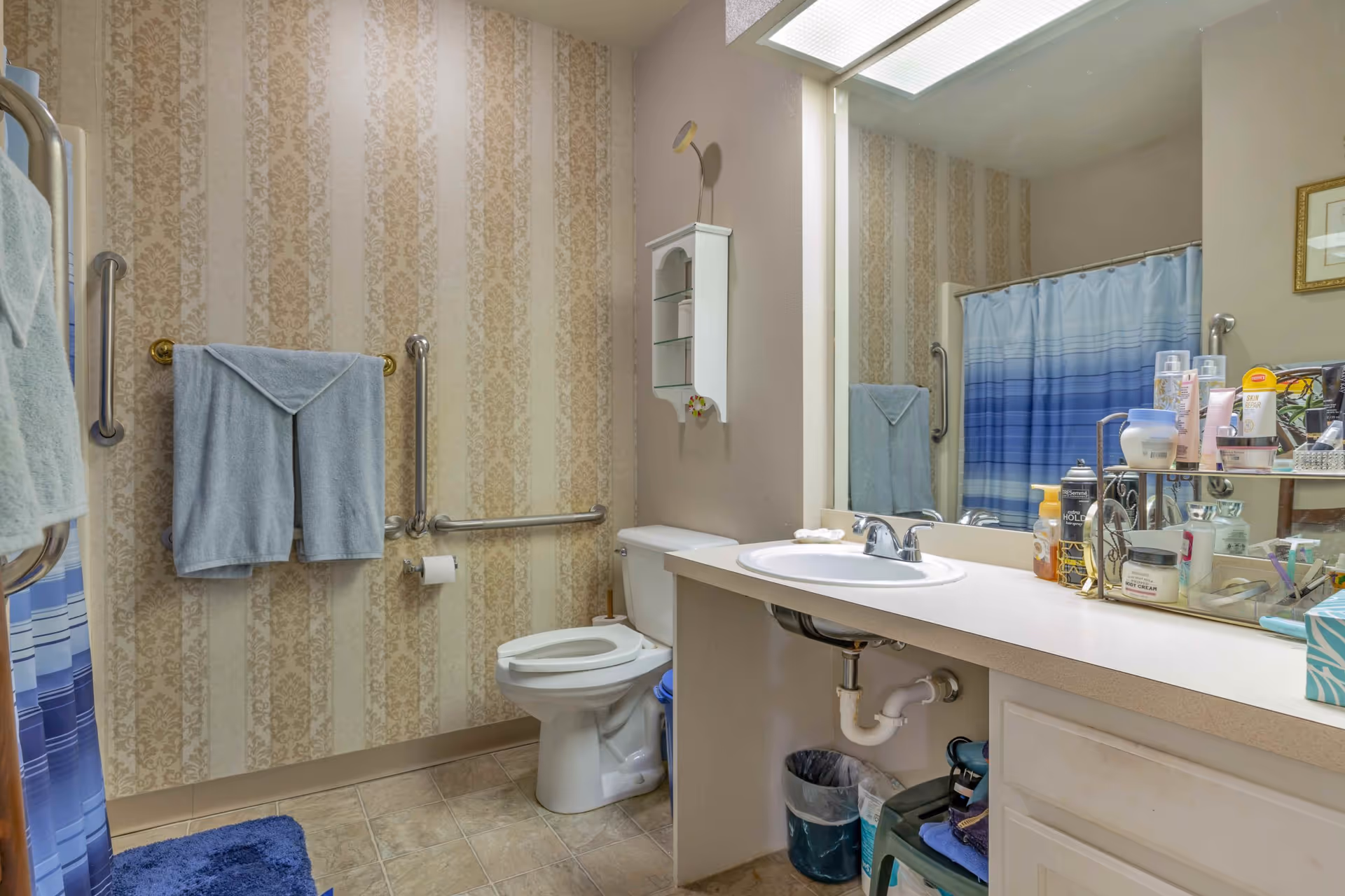 A bathroom with beige patterned wallpaper, a white toilet with grab bars around it, a white sink with a large mirror above it, and a countertop filled with various toiletries. There is a blue and white striped shower curtain and blue towels hanging on the wall. The floor is tiled and there is a blue bath mat on the floor.