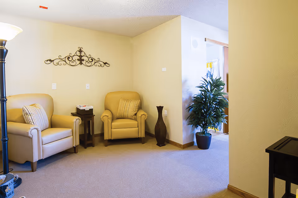 A cozy living room area with two beige armchairs, each with a striped cushion, a small wooden side table between them holding a basket, a tall floor lamp on the left, a decorative wall piece above the chairs, a tall dark vase on the floor, and a potted plant near an open doorway leading to another room.