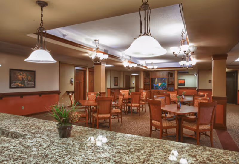 Interior view of a dining room in an assisted living facility with multiple wooden tables and chairs arranged neatly. The room is warmly lit with hanging pendant lights and chandeliers. There is a granite countertop in the foreground with a small potted plant on it. The walls are decorated with framed artwork and the carpeted floor adds to the cozy atmosphere.